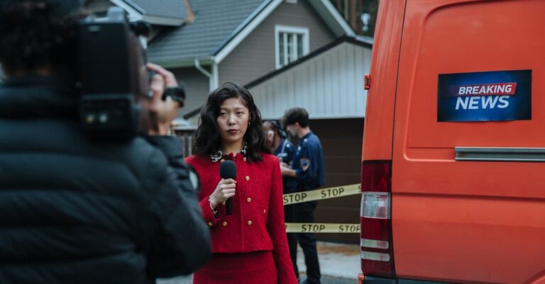 Female reporter in red with microphone reporting live at an outdoor crime scene with a news van.
