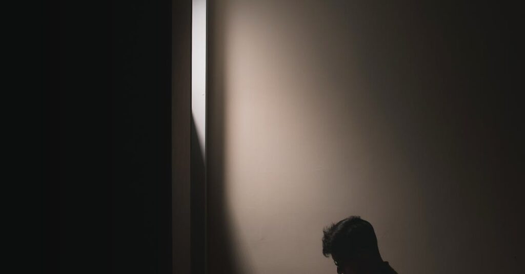 Silhouette of a man sitting on a bed in a dark room, reading a book by window light.