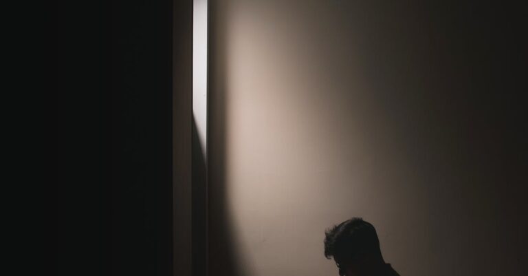 Silhouette of a man sitting on a bed in a dark room, reading a book by window light.