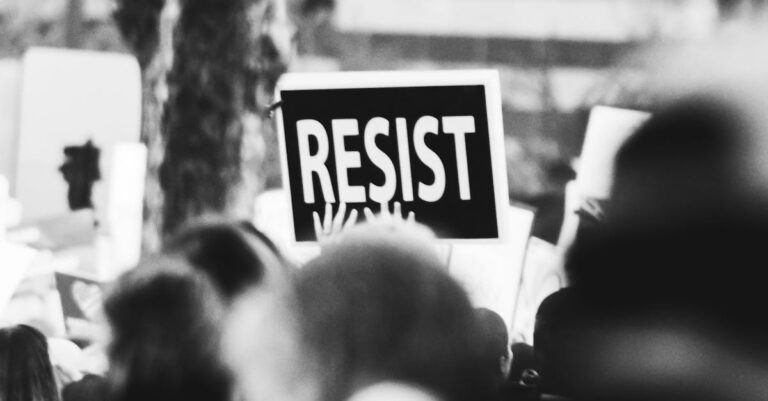 Monochrome image showing a crowd holding a 'Resist' sign at a protest.