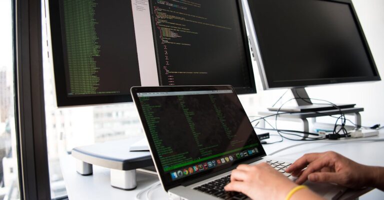 A developer writes code on a laptop in front of multiple monitors in an office setting.