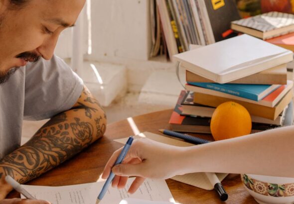 A tattooed Asian student concentrates on studying with books and papers at a desk.