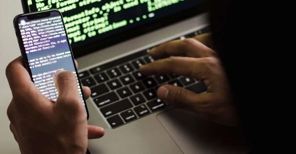 A hooded figure engaged in hacking using a laptop and smartphone in low light.