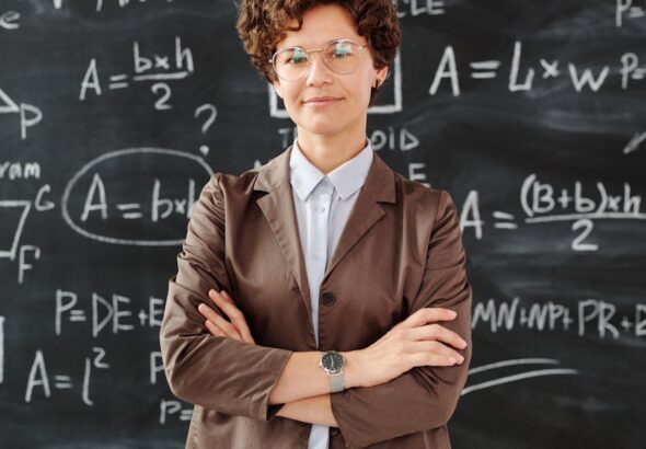 Confident female teacher standing with arms crossed in front of a detailed mathematical blackboard.