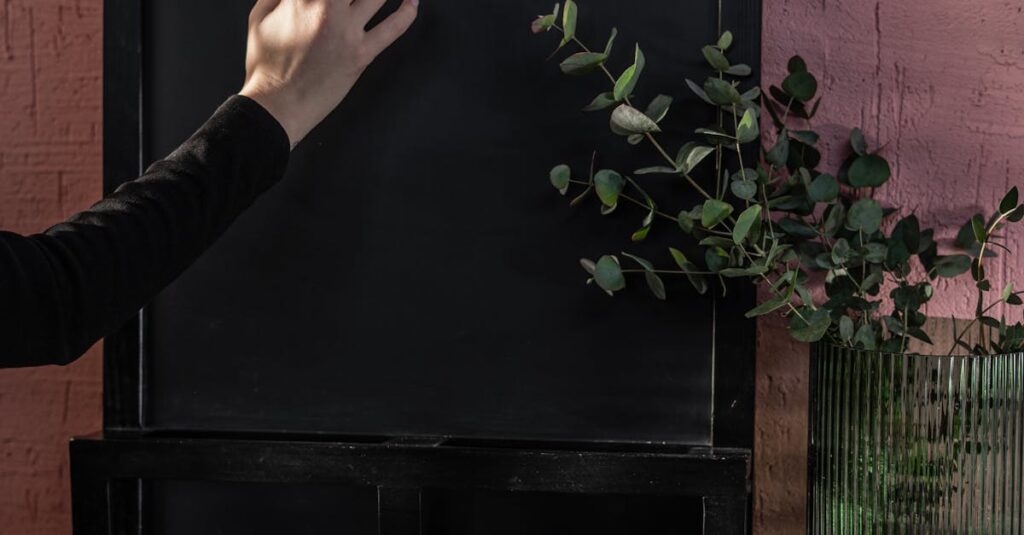 A hand writing with chalk on a blackboard, accompanied by a green plant, set indoors.