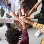 A diverse group of professionals joins hands in a team-building exercise indoors.