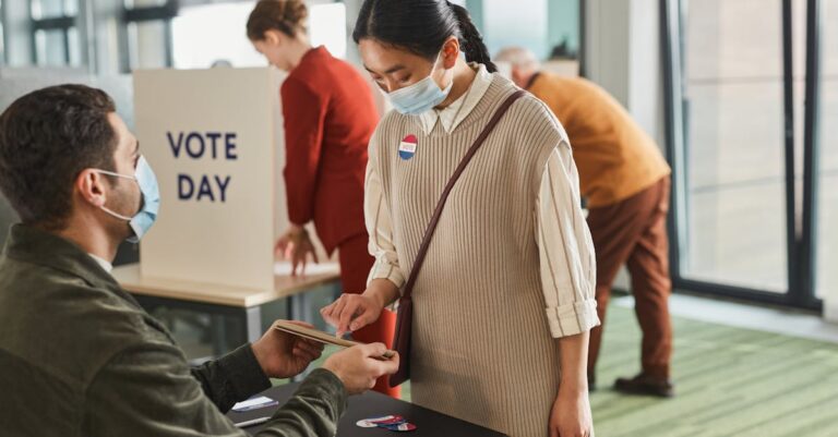 Voters casting ballots indoors on Election Day, wearing masks and using tablets for contactless voting.