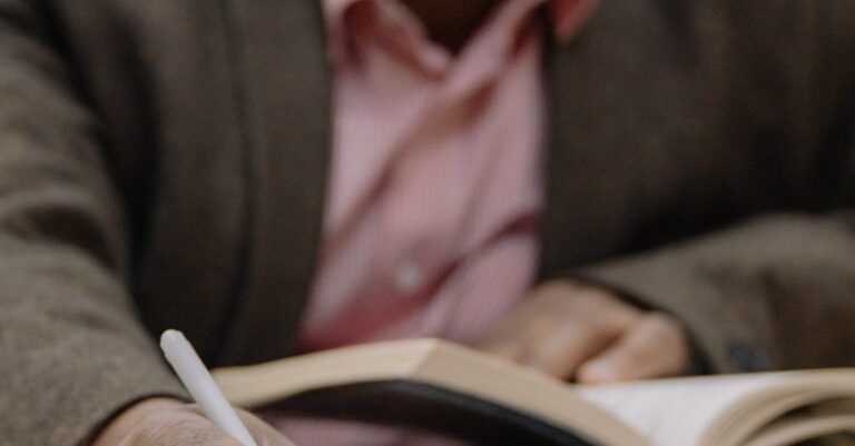 Adult man studying in a library, writing notes from an open book, with focus on hand and pen.