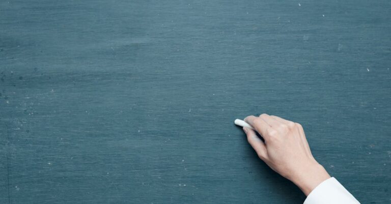 Hand holding chalk ready to write on a blank blackboard surface.