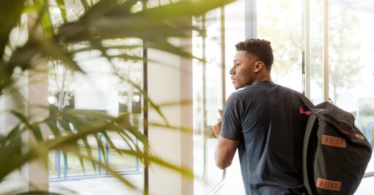 A college student with a backpack exits a campus building through glass doors.