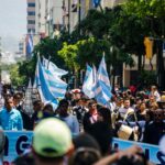 A lively urban protest with people holding flags and signs on a sunny day.