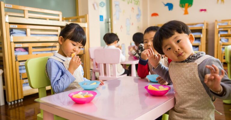 Kids seated around a table in a colorful classroom, eating snacks happily.
