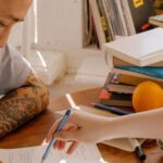 A tattooed Asian student concentrates on studying with books and papers at a desk.