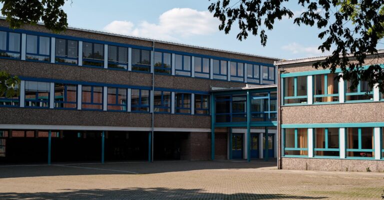 Exterior view of a modern educational building in Geilenkirchen, Germany, featuring large windows and a paved courtyard.