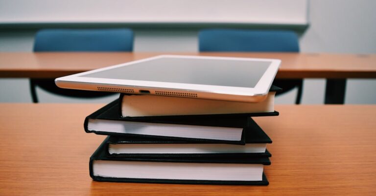 A tablet rests on top of a stack of books in an empty classroom, illustrating modern education.
