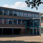 Exterior view of a modern educational building in Geilenkirchen, Germany, featuring large windows and a paved courtyard.