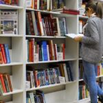 A young woman stands reading a book in a well-stocked library.