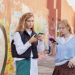Two fashionable teenagers using smartphones in an urban graffiti setting, showcasing modern youth culture.