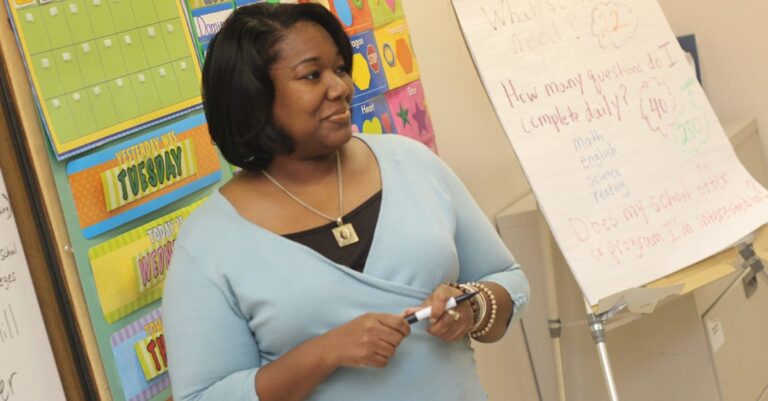 A female teacher standing by a whiteboard in a colorful classroom setting.
