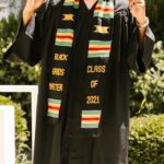 Smiling graduate wearing cap and gown celebrating outdoors with finger point gesture and colorful stole.