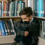 Young boy in library appears sad, sitting alone among bookshelves, suggesting themes of solitude and bullying.