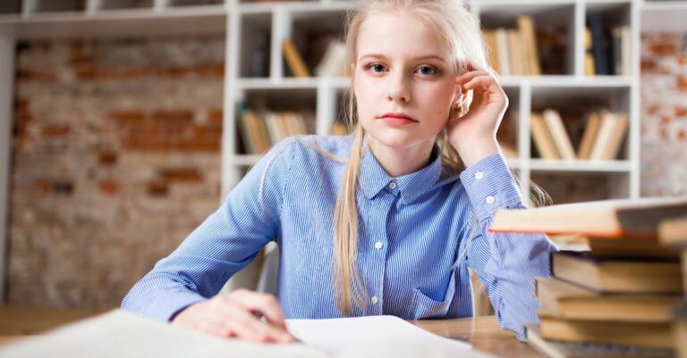 A young woman with blond hair studying at a table piled with books.