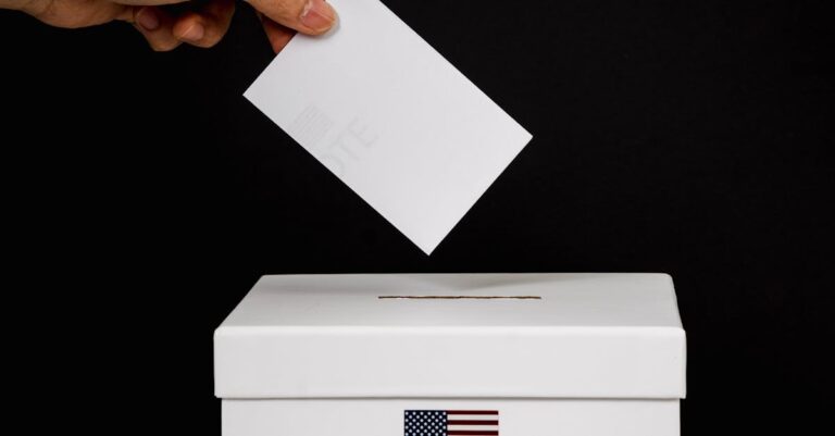 Close-up of a hand placing a ballot into a voting box marked 'Vote' with a USA flag.
