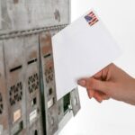 Close-up of a hand inserting a stamped envelope into a mailbox, symbolizing voting.