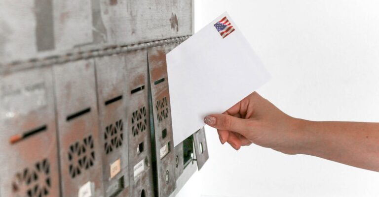 Close-up of a hand inserting a stamped envelope into a mailbox, symbolizing voting.
