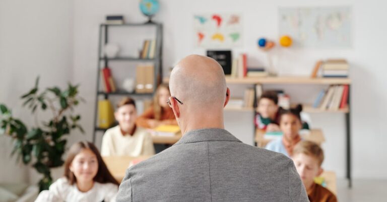 Bald teacher in a classroom engaging with attentive students sitting at desks.