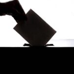 Silhouette of a hand placing a vote into a ballot box, symbolizing democracy.