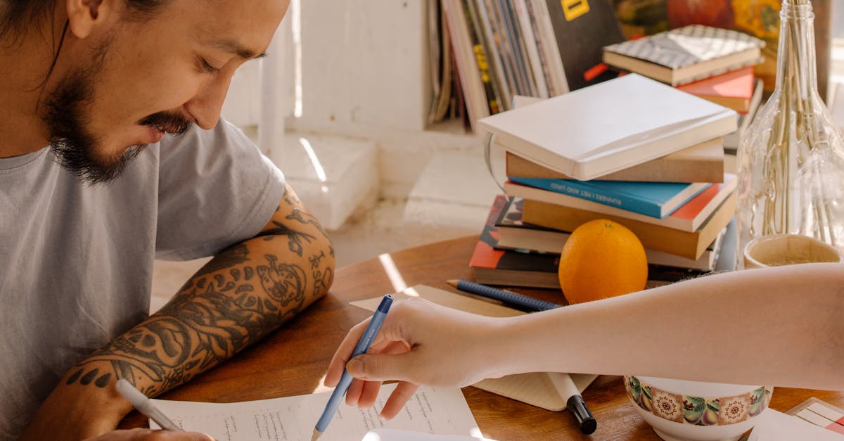 A tattooed Asian student concentrates on studying with books and papers at a desk.