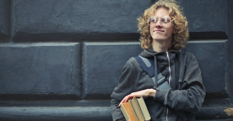 A smiling young man with glasses and messy hair holds books, leaning against a textured wall.