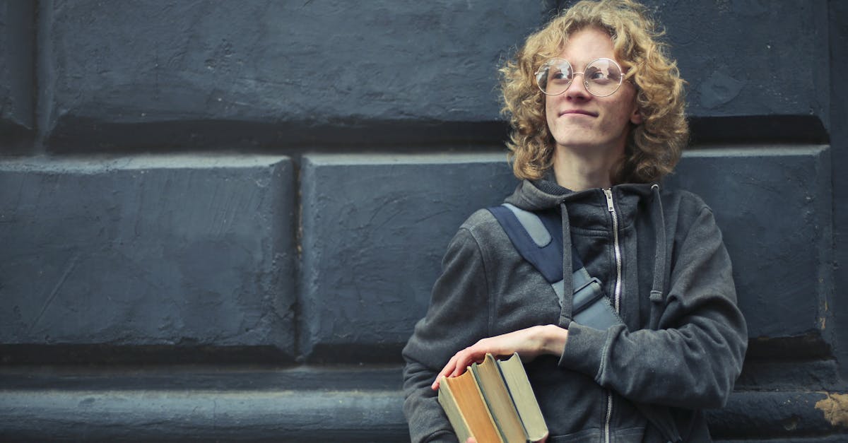 A smiling young man with glasses and messy hair holds books, leaning against a textured wall.