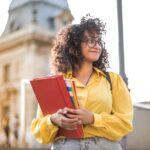 Smiling female student with curly hair and glasses holding books on campus steps.