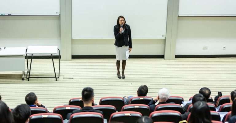 Female speaker presenting to an audience in a modern auditorium setting.