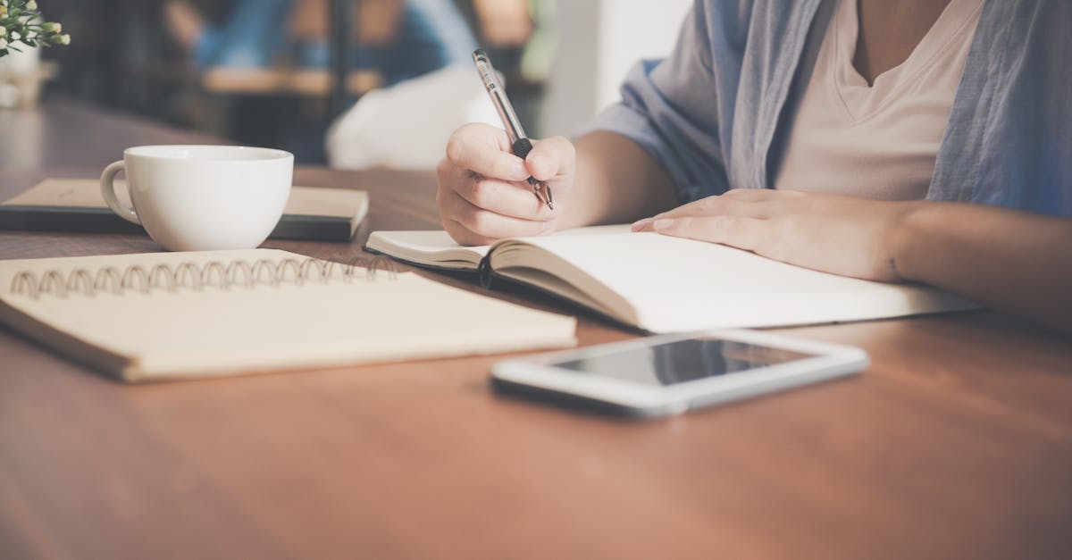 A woman writes in a notebook at a café table with a coffee and smartphone nearby.
